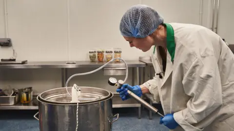 Bwblin A woman in a laboratory white coat checks a pressure gauge attached to a steel vat inside a food making facility. She is wearing a blue hairnet and blue plastic gloves. 