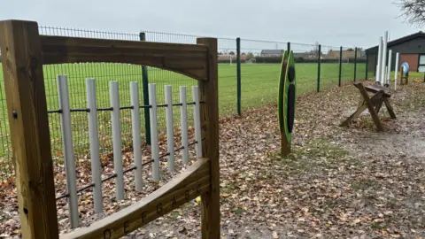 The image shows an outdoor musical play area set along a fenced boundary of a grassy field. 