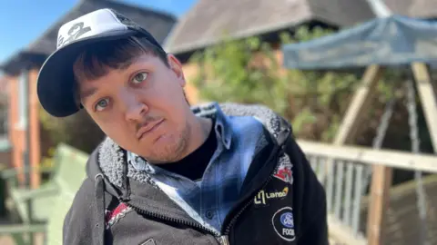 A man with a hat on looks at the camera. He is wearing a black T-shirt, blue plaid shirt and a black zip-up hoodie. The sun is shining and there are benches and bushes in the background. 