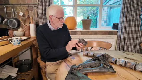 A white haired man wearing a blue jumper examines a dinosaur bone in his hands while sitting in his kitchen at the table 