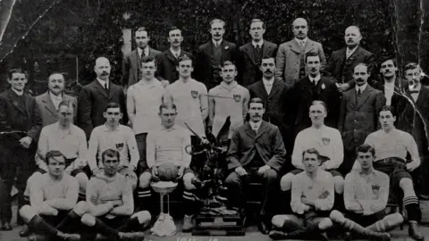 E Gill/Neil Harvey A black and white photograph from 1912 showing 27 men sitting and standing in rows. Many of the men are dressed in suits with waistcoats, but others are dressed in football kit, and one is holding a football. There is  large (2ft/0.6m) trophy showing a winged male figure holding a laurel leaf and trampling on a figure below