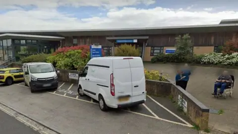 A single-storey hospital building with a number of vehicles parked outside and some anonymous people.