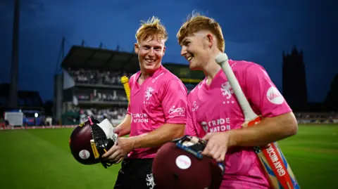 Getty Images Two Somerset cricketers in pink kit smile as they walk off the pitch at Taunton after a T20 match. It is after dark and the pitch and stadium are lit up by floodlights. A large church tower and viewing stand are silhouetted in the background.