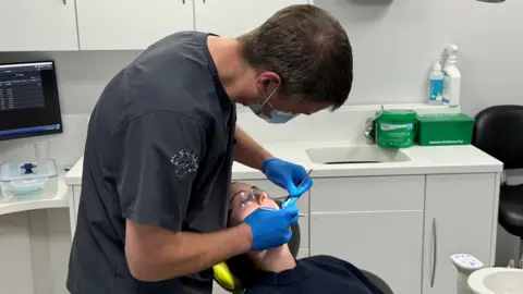 Dr Russell Gidney is leaning over a female patient who is reclined in the dental chair. he is using two tools to look in her mouth. the background shows other dental equipment including a side sink and computer.