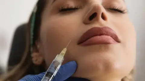 A woman with long brown hair has a needle injected into her face in a close-up photo of her face. She has make up on and the needle is near her mouth. The person injecting it has blue gloves on.