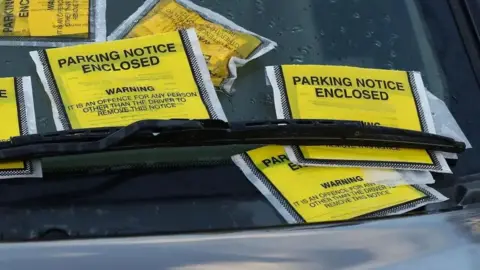 A stock image of six yellow parking enforcement tickets stuffed under a windscreen wiper on the front of a car