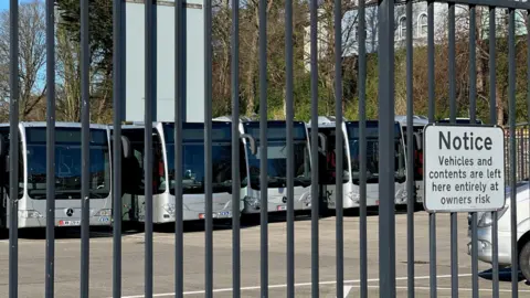 A row of silver single decker buses behind a fence at the Bus Vannin depot.