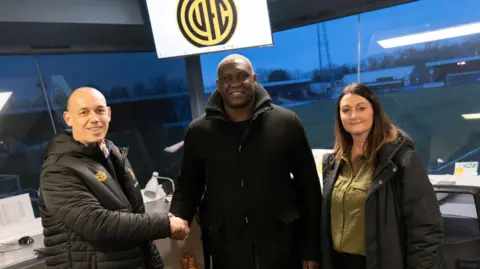 FootballSafetyApp Emile Heskey stands between Rich Cooksley and Sarah King. He is shaking Cooksley's hand. They are in the offices of Cambridge United and the ground can be seen clearly through large windows.