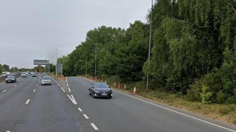 A google maps image of cars on a major A road showing a slip road to the right and large trees along the roadside 