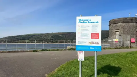 Cheddar Reservoir seen from a footpath next to it. There is a sign saying 'Welcome to Cheddar Reservoir' in the foreground of the photo. The Mendip Hills are in the background.