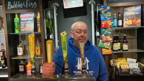 Dave Owen stood behind a bar wearing a blue hoodie with white writing on it. He is bald and is holding onto one of the beer taps.