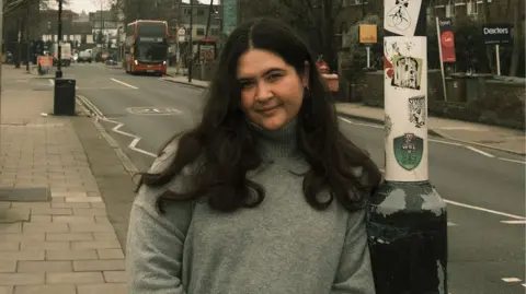 Dani Gonçalves A woman with long black hair wears a light grey woolen sweater as she leans against a lamp-post. A London street stretches out behind her, and a double-decker red bus approaches on the opposite side of the road.