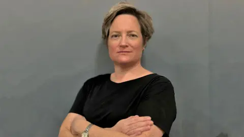 Claire Stuckey Claire Stuckey standing against a dark grey background with her arms folded. She is looking at the camera with a determined expression. She is wearing a black t-shirt, a silver watch, and has short brown hair in a side parting.