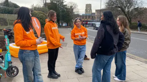 Three women dressed in orange hoodies with the word Enough on them talking to two female students in Bristol.