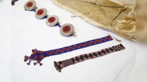 David McHugh The image shows three artefacts that look like jewellery lying on a white table.