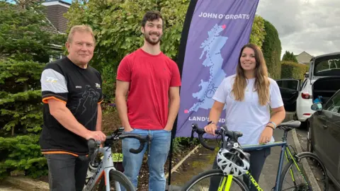 Two men and a woman with bicycles next to a flag showing the route from Land's End to John O'Groats