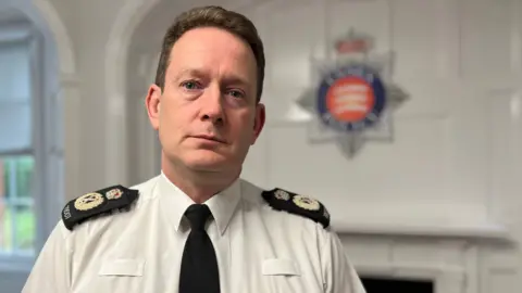Stuart Woodward/BBC A policeman wearing a white shirt and a black tie with black epaulettes. He is standing in front of a white wall with an Essex Police crest in the background.