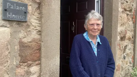 Sue Massey is a woman of 80 wearing a purply blue cardigan over a lighter blue floral shirt. She is standing at the doorway of her home which is a stone building with a deep dark burgundy-brown front door. A grey slate sign to the side of the door carries the name Palace House.