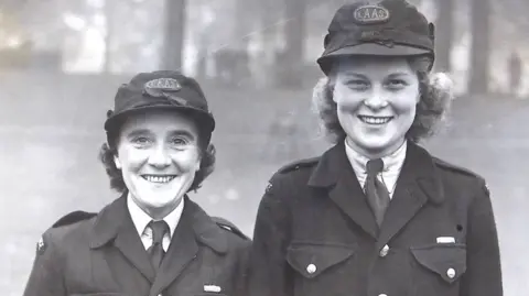 London Ambulance Service N Armitage and Betty Leorton saw the camera. Anne has black, short hair and wears a hat. Betty is long and turns golden hair and wears a hat.