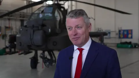 Luke Pollard MP wearing a blue suit and a red tie and talking in front of an Apache helicopter.