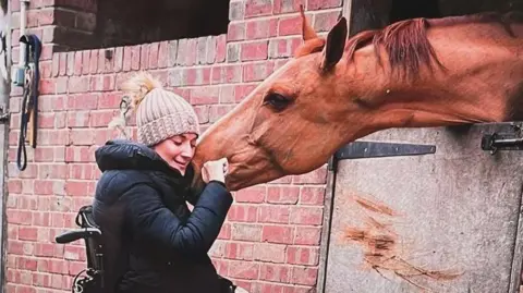 SARRA WILSON Sarra Wilson sits in her wheelchair hugging a horse in her stables. The horse is a rich tan colour and is sticking his head above the stable door. Mrs Wilson is wearing a brown wool bobble hat and a black coat. She has her eyes closed and her hand is stroking the nose of the horse.