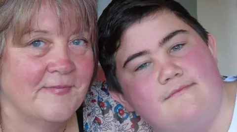 A woman with short blonde hair, wearing a floral-patterned dress, next to a young man with short black hair and wearing a white t-shirt.