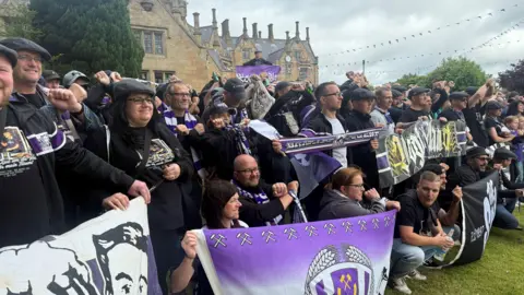 A large group of Erzgebirge Aue fans holding flags and banners standing outside Brownlow House - a brown brick building in a Baronial style
