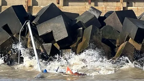 Yacht sinking in water near large black rocks. Only the mast is visible.