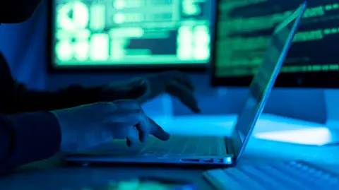 A stock image of a person at a laptop with several screens in the background displaying numbers and code.