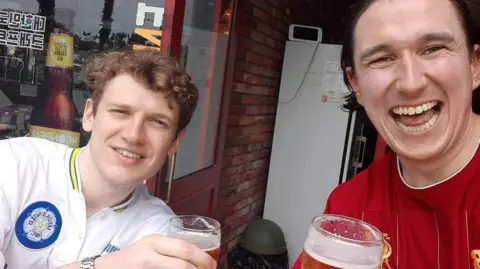 Josh Wileman Two football fans, one in a Leeds United shirt and one in a Liverpool shirt, pose with pints of beer in hand.