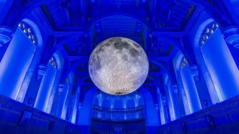 The large grey sculpture of the Moon hanging in a cathedral which is lit up with blue lights. It hangs from the ceiling and is surrounded by the interior pillars of the building.