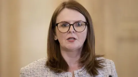 Caoimhe Archibald in white tweed jacket and black top. She has red hair and brown glasses.