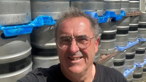 Martin Brunnschweiler smiles, he has short grey hair and wears a dark T-shirt and silver-rimmed glasses. He stands in front of silver kegs of beer which have been stacked up on top of each other.