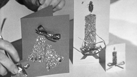 Black and white image of Valerie's hands behind 3 Christmas cards. One on left is dark card with two shiny bells and a bow, and then on the right, two light coloured cards, one tall one and one little one, with candles on them.