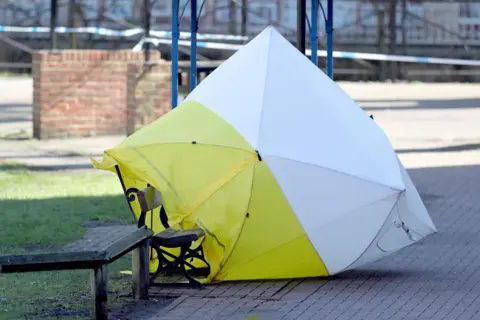 PA Media A yellow and white forensic tent covers a bench in front of a patch of grass. Police tape is visible in the background. 