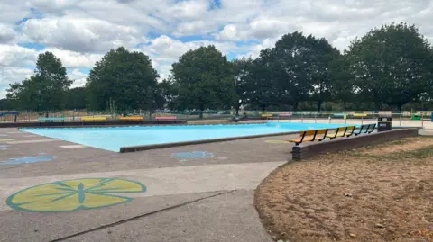 A drained paddling pool is pictured surrounded by parched grass. There are brightly coloured benches and colourful drawings on the concrete around the pool.