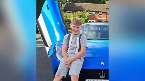 Family photograph Cole, 11, with blond hair wearing a grey and white t-shirt and shorts set. He is perched on a blue sports car with the drivers door open parked on a housing estate. It is a sunny day. Cole is smiling.