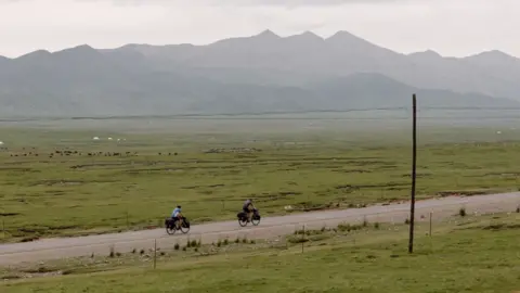 Kohler family Two cyclists ride left to right across the image along a straight road through a green flat landscape, with mountains rising in the background 