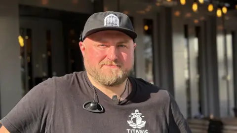 Simon Thake/BBC A pub worker wearing a black t-shirt and black baseball cap. He has fair-coloured beard.
