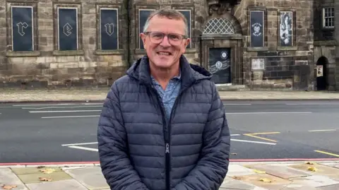 Jon Plant, a man wearing glasses, as well as a blue shirt and a dark-coloured jacket, is standing by the side of a road. He is smiling for the camera. There is a stone building in the background.