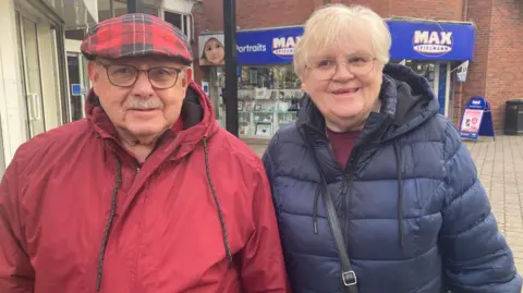 Mr and Mrs Lees in Crewe town centre. He has a moustache and is wearing a red-check flat cap and a red raincoat. She is dressed in a blue puffer-style coat and is also wearing glasses and a handbag strapped over her right shoulder.