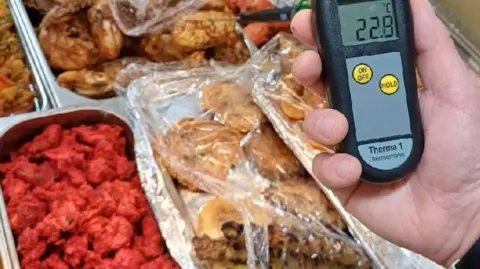 Red curry and assorted brown meats being stored in open stainless steel containers. Some is covered in cling film. A close-up of a hand on the right of the picture shows a thermometer with a reading of 22.9C. 