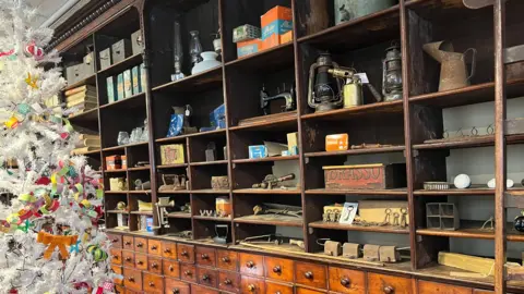 John Devine/BBC Mid-19th Century shelving in dark wood, possibly mahogany.
There are several rows of small drawers underneath the shelving, and lots of old-fashioned items on the shelves.