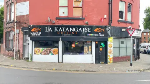 The front of the cafe, with the words 'La Katangaise' in white letters on a black background on its sign. There are two orange-coloured fish on each side of the sign. The red-brick building is next to a road, which curves round in front of it.