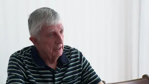 John Benson sitting down and looking at someone out of frame. He is wearing a T-shirt with green, dark blue and white stripes.