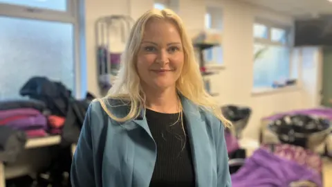 BBC / ELIZABETH BAINES A woman with platinum blonde hair wearing a pale blue blazer and black top. Behind her many towels indicate the salon where young people can enjoy hair styling.