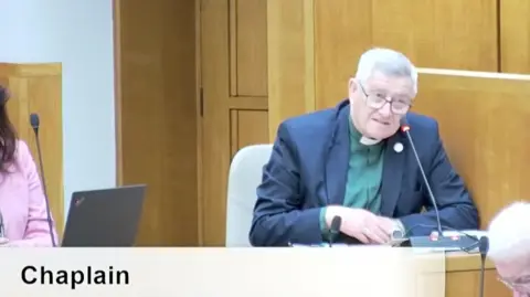 Norfolk County Council Albert Cadmore sits in the council chamber wearing a green shirt, blue jacket and clerical dog collar. Two people near by have their heads bowed.