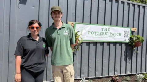 A woman wearing a black polo shirt and a man wearing a green polo shirt are standing next to a grey shipping container which has a white banner on with green lettering which promotes The Potters Patch community garden. There is a hanging basket on each end of the banner attached to a wooden plank.