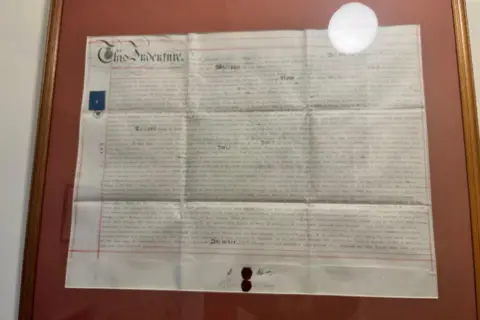A framed white paper document dense with writing, mounted on maroon card with a wooden frame. In the top corner, decorative writing reads "This Indenture."