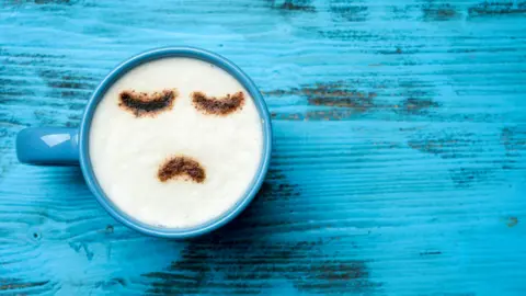 A birds-eye view of a blue coffee mug containing a cappuccino. The frothy top is dusted with chocolate in a sad emoji design. The mug sits atop a wooden table painted in a similar cool blue colour.